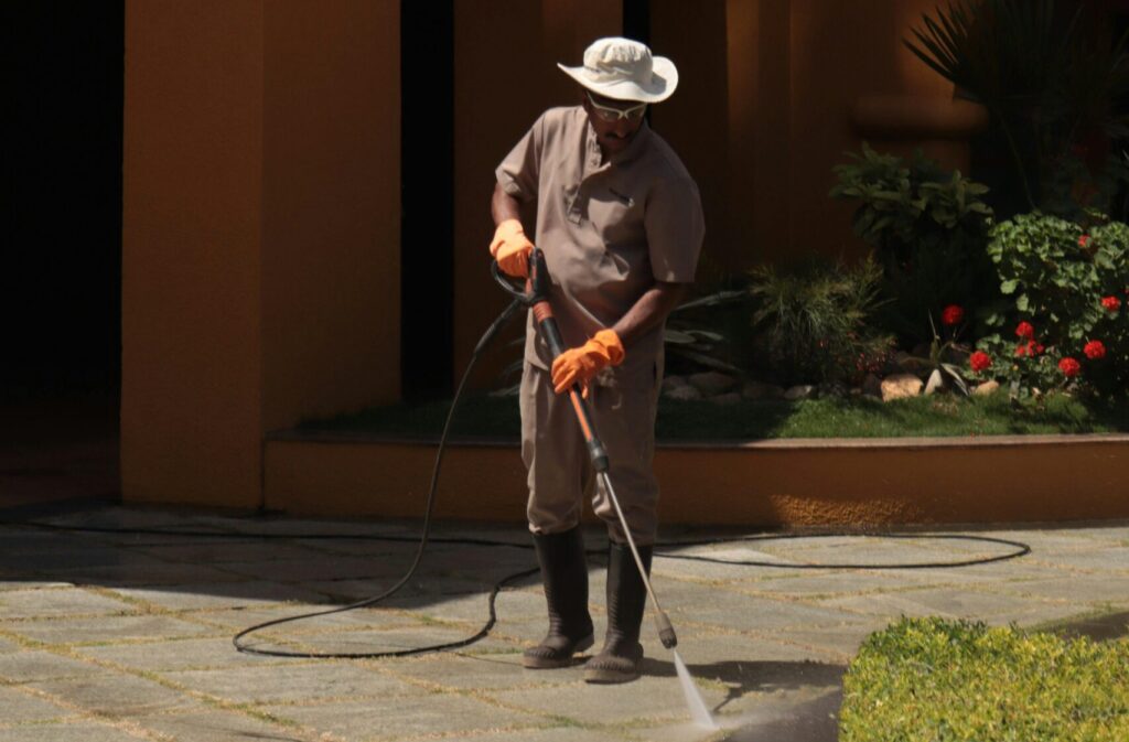 A gardener wearing a hat cleans an urban sidewalk with a pressure washer.